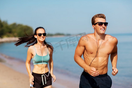 培训科技摄影照片_健身、运动和科技概念-戴着耳机在夏日海滩跑步的幸福情侣。戴着耳机在海滩上奔跑