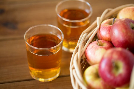 木桌子摄影照片_水果，食物和收获概念—在柳条篮和玻璃果汁在木桌上。篮子里的苹果和桌子上的果汁杯