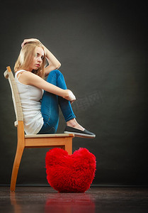 心碎的爱情观。悲伤不快乐的女人坐在椅子上，地板上的红心枕头，黑暗的背景
