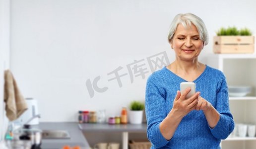 技术和老年人概念-微笑的老年女性使用智能手机而不是厨房的背景。微笑的老妇人在厨房里使用智能手机