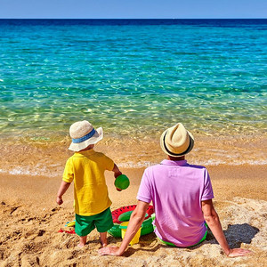 孩子坐爸爸背上摄影照片_两岁的学步男孩和父亲在海滩上。充气戒指和沙滩玩具。暑假全家度假。希腊西索尼亚。