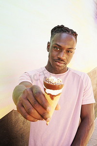 黑色t恤摄影照片_一个年轻英俊的黑人男子穿着粉红色的T恤，在夏天的日出或晴天的墙上拿着一个冰淇淋吃