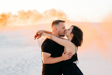男孩和一个女孩在黑色的衣服拥抱和运行在白色的沙滩上与橙色的烟雾