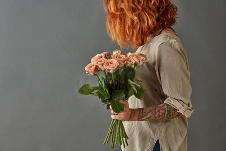 女孩拿花摄影照片_年轻的红头发女孩与纹身持有一束粉红色的玫瑰在灰色背景。一个年轻的女孩拿着一束粉红色的花
