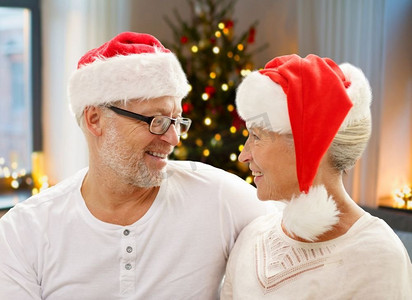 圣诞节老人摄影照片_节日、圣诞节和人们概念快乐的老年夫妇在家中戴着圣诞老人的帮手帽子。圣诞快乐的高年级夫妇在家中
