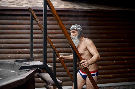 滑稽的老头爬楼梯到桑拿房外的木头浴缸。温泉度假村和热水浴。老年男子爬楼梯到外面的浴缸