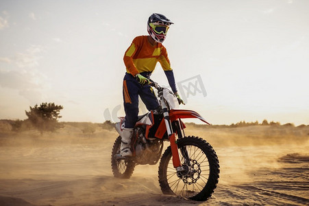 沙漠中的越野摩托车手戴着防护头盔，穿着防护服骑着运动摩托车。骑在运动摩托上的摩托车手在尘土飞扬的风景中