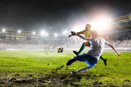 足球运动员在夜间体育场与燃烧的灯踢足球。两个足球运动员在体育场的行动