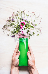 持有绿色化妆品产品瓶子的女性手与植物和花，顶视图的白色背景天然化妆品概念