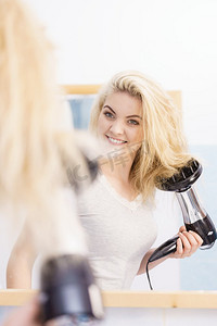 发型设计摄影照片_积极的女人使用吹风机在她的金发发型。头发护理，发型设计概念..金发女郎的女人使用吹风机