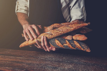 新鲜出炉的长棍面包拿着一个男人的手对着一张木桌的背景，色调照片。’男人的手握着长棍面包’