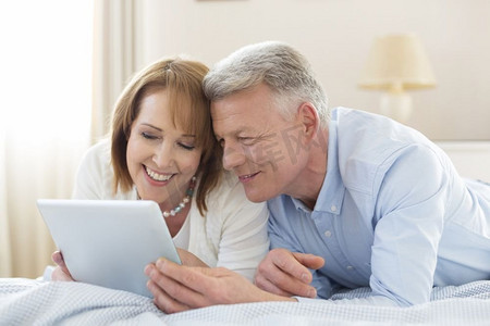 微笑的成熟夫妇在家躺在床上分享数字平板电脑
