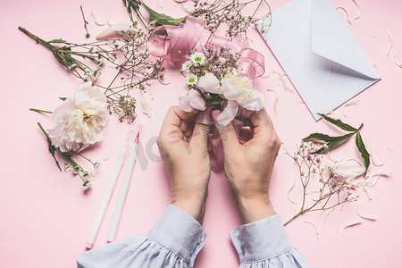 女性手做美丽的花安排与牡丹花在柔和的粉红色背景与信封，顶视图