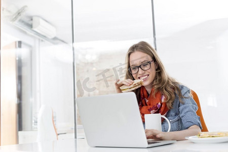富有创意的女商人一边在办公室使用笔记本电脑一边吃早餐