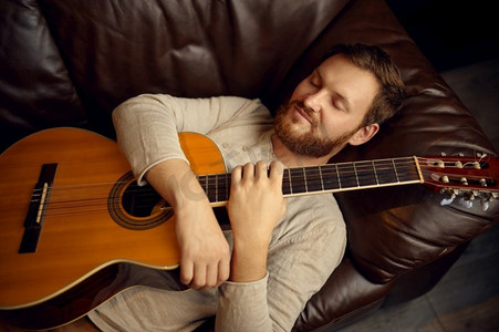 年轻人躺在沙发上拥抱吉他。吉他手的激情和乐趣。音乐家在家休息。年轻人躺在沙发上吉他