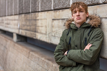 抑郁的孩子摄影照片_年轻的成年男性青少年男孩外面在一个城市城市穿着一件绿色皮大衣靠在墙上