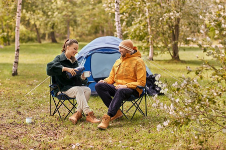女人倒茶摄影照片_露营、旅游和旅行概念—愉快的夫妇与热水瓶倒茶杯在帐篷营地。快乐的夫妇与热水瓶倒茶在帐篷营地