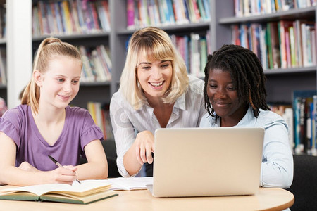 女高中生摄影照片_老师帮助两名女高中生在图书馆用笔记本电脑工作
