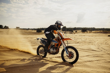 摩托车宝马水鸟摄影照片_越野摩托车手在沙丘上行驶，越野自行车从车轮下吹起灰尘。专业越野摩托车手在沙丘上驾驶