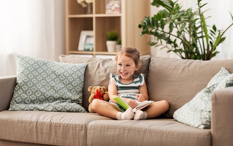 童年和人的概念—快乐的小女孩坐在沙发上与书和玩具泰迪熊在家里。快乐的女孩坐在沙发与书在家里