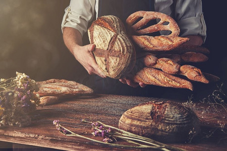食物调色摄影照片_面包师手里拿着面包，一张木质棕色桌子，黑色背景上有干花，调色照片。各式各样的面包手&S手中
