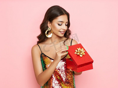 漂亮的女人在一个鸡尾酒礼服打开礼物盒。美丽的模特与长卷发。圣诞节或新年庆祝活动。节日与聚会