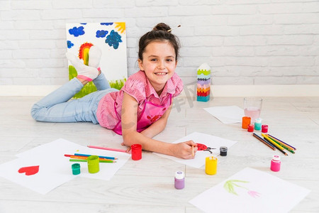 创意裂纹笔刷摄影照片_微笑的小女孩躺在地板上画油漆刷白纸