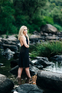蓝色的裙子摄影照片_金发女孩在一件黑色的裙子与蓝色的眼睛在河的石头海岸