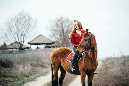 一个穿着红色羊毛衫和黑色高靴的红发骑师女孩，骑着一匹马散步