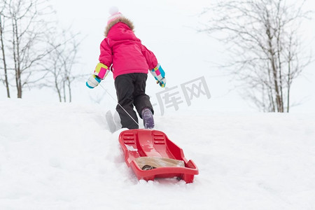爬雪山摄影照片_童年，雪橇和季节概念-快乐的小女孩，冬天拉着雪橇爬雪山。冬天雪山上拉雪橇的小女孩