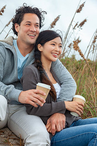 年轻的亚裔华人男女，男孩女孩，坐在海滩上微笑着，牙齿完美的夫妇，喝着外卖茶杯或咖啡