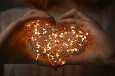 手写圣诞艺术字体摄影照片_女性的手在一个心脏的形式举行圣诞灯串在黑暗的背景。女人的手在黑暗中握着一串灯。