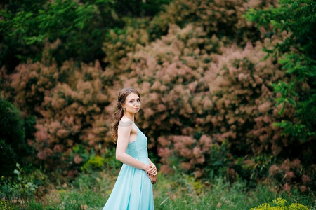 花园玫瑰摄影照片_一件绿松石长连衣裙的快乐女孩在一个绿色公园在草本植物、树和玫瑰灌木丛的背景