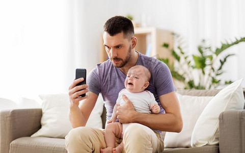 家庭，为人父母和技术概念—父亲与哭泣的小婴儿和智能手机在家里。父亲与哭泣的婴儿和智能手机在家里