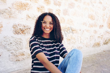一幅快乐微笑的年轻黑人女子的肖像，戴着眼镜，牛仔裤和条纹T恤，坐在地上，留着卷发