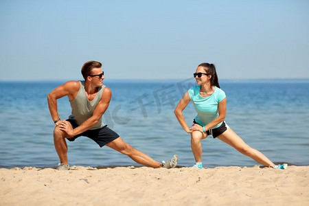 健身、运动和生活方式理念--训练前微笑的情侣在海滩上伸展双腿。微笑的情侣在海滩上伸展双腿