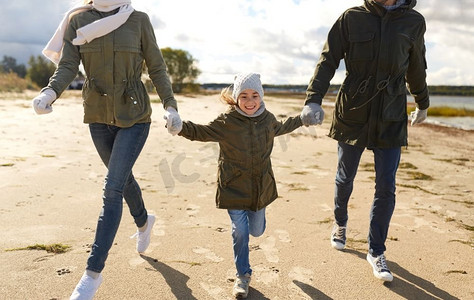家庭、休闲、人的概念-快乐的父母和小女儿沿着秋天的海滩奔跑。幸福的一家人沿着秋天的海滩跑步