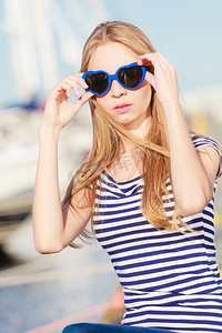 心形太阳镜摄影照片_夏天和人们的概念时尚可爱的金发女郎与心形太阳镜在码头反对游艇在港口