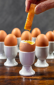 人浸入烤面包士兵进入一组鸡蛋的一个煮鸡蛋在一个木桌上的白色鸡蛋杯 