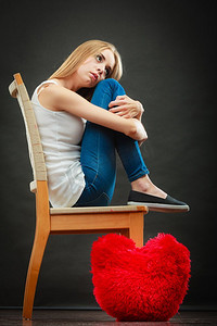 心碎的爱情概念。悲伤的不愉快的妇女坐在椅子红色的心枕头在地板黑暗的背景