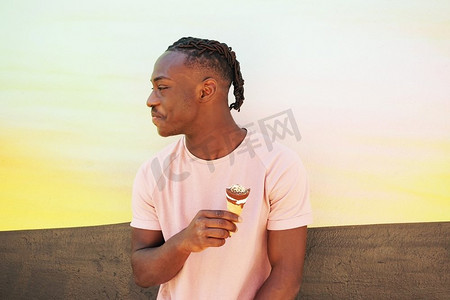 黑色t恤摄影照片_一个年轻英俊的黑人男子穿着粉红色的T恤，在夏天的日出或晴天的墙上拿着一个冰淇淋吃