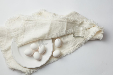 鸡生蛋摄影照片_白色餐桌上餐巾上盘子里的生鸡蛋俯瞰。生鸡蛋