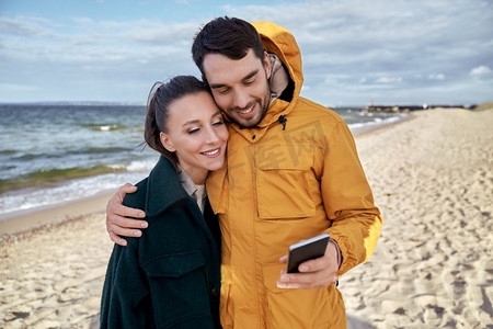 技术，关系和人的概念—快乐的夫妇与智能手机在秋天的海滩。夫妇与智能手机在秋季海滩