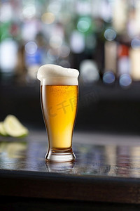 一杯啤酒与泡沫在黑木的一张桌子在酒吧在模糊的酒吧背景。玻璃啤酒在木桌和模糊的背景。