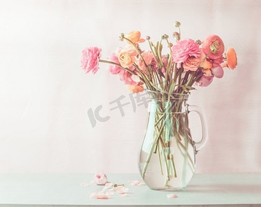 粉彩粉红色毛茛花花束在玻璃壶在桌子上，前视图