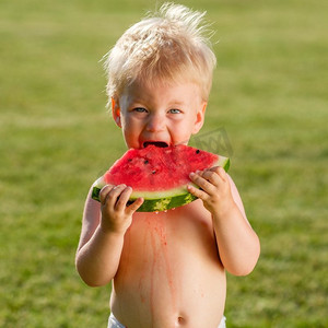 吃西瓜场景摄影照片_户外学步儿童的肖像。一岁男婴在花园里吃西瓜片的乡间场景。快乐孩子脏兮兮的脸。