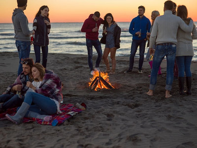 年轻夫妇坐在周围篝火在海滩上的朋友在日落喝啤酒。夫妇享受与朋友在日落在海滩