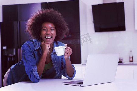 年轻面带微笑的黑人妇女在现代厨房里一边玩电脑，一边喝咖啡