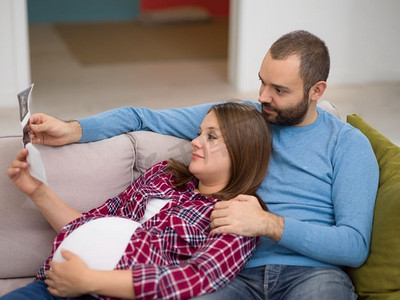 怀孕的夫妇寻找婴儿的超声波。’年轻的怀孕夫妇寻找婴儿的超声波照片，而放松在沙发上在家里’