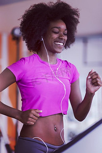 一名非裔美国女性在健身房的跑步机上跑步，同时戴着耳机听音乐。在跑步机上跑步的美国黑人妇女